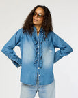 A person wearing sunglasses and the Kerri Rosenthal Molly Denim Shirt with ruffle trim, paired with lightweight denim jeans, confidently stands with hands on hips against a plain light background.