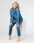 Wearing a Molly Denim Shirt by Kerri Rosenthal, featuring a ruffle trim, and lightweight denim jeans, a barefoot individual sits on a white rectangular block against a stark white background. Their long hair cascades down as they gaze slightly upwards, hands gently resting on their knees.