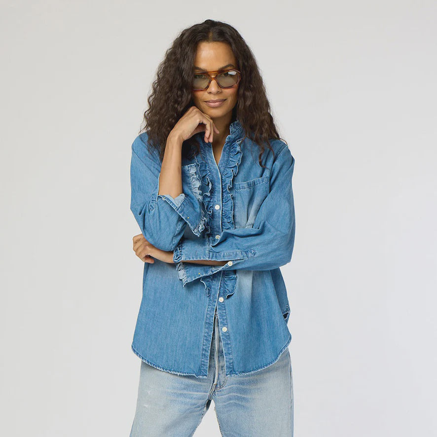 Wearing a Molly Denim Shirt by Kerri Rosenthal, a person with long, wavy hair confidently poses with crossed arms. The shirt's ruffle trim pairs well with their light-colored jeans against a plain backdrop, and stylish sunglasses add the finishing touch to the look.