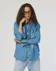 Wearing a Molly Denim Shirt by Kerri Rosenthal, a person with long, wavy hair confidently poses with crossed arms. The shirt's ruffle trim pairs well with their light-colored jeans against a plain backdrop, and stylish sunglasses add the finishing touch to the look.