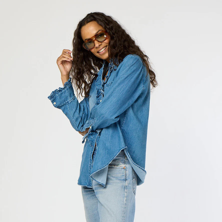 A person wearing sunglasses and a Kerri Rosenthal Molly Denim Shirt with ruffle trim poses confidently against a plain background, paired with lightweight denim jeans.