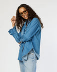 A person wearing sunglasses and a Kerri Rosenthal Molly Denim Shirt with ruffle trim poses confidently against a plain background, paired with lightweight denim jeans.