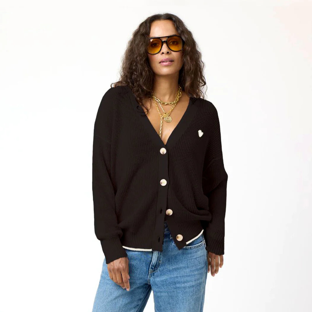 A woman with long curly hair, wearing large orange sunglasses and layered gold necklaces, poses confidently. She is dressed in a black Kerri Rosenthal Slouchy Cardigan with a heart appliqué over a white top and blue jeans. The background is plain white.