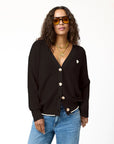 A woman with long curly hair, wearing large orange sunglasses and layered gold necklaces, poses confidently. She is dressed in a black Kerri Rosenthal Slouchy Cardigan with a heart appliqué over a white top and blue jeans. The background is plain white.