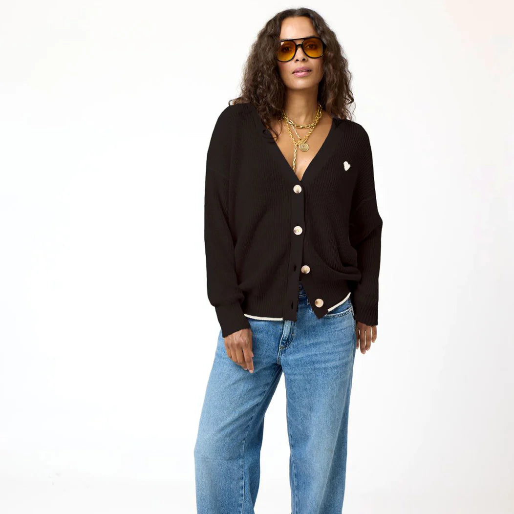 A person with long, curly hair wears yellow-tinted sunglasses and a Kerri Rosenthal Slouchy Cardigan featuring a white heart appliqué. Layered gold necklaces adorn their neck over a white undershirt, paired with blue jeans. They stand against a plain white background, casually posing with one hand by their side.