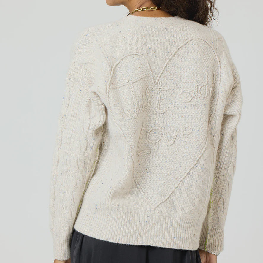 A person dressed in the Suzanne Just Add Love Cashmere Cardigan by Kerri Rosenthal, a cream-colored knitted sweater crafted from Donegal Cashmere, featuring a large heart design on the back with the words "Just add Love" stitched inside. The cardigan beautifully showcases textured cable patterns on the sleeves.