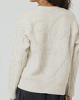 A person dressed in the Suzanne Just Add Love Cashmere Cardigan by Kerri Rosenthal, a cream-colored knitted sweater crafted from Donegal Cashmere, featuring a large heart design on the back with the words "Just add Love" stitched inside. The cardigan beautifully showcases textured cable patterns on the sleeves.