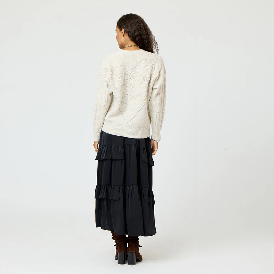 A person with long curly hair is standing with their back to the camera, wrapped in a luxurious Suzanne Just Add Love Cashmere Cardigan by Kerri Rosenthal and a black tiered skirt, paired with brown boots. The soft elegance of the outfit contrasts subtly against the plain gray background.