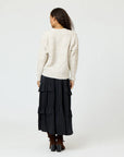 A person with long curly hair is standing with their back to the camera, wrapped in a luxurious Suzanne Just Add Love Cashmere Cardigan by Kerri Rosenthal and a black tiered skirt, paired with brown boots. The soft elegance of the outfit contrasts subtly against the plain gray background.