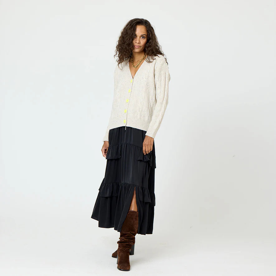 A woman with curly hair is wearing a Suzanne Just Add Love Cashmere Cardigan by Kerri Rosenthal, featuring yellow buttons, over a long, tiered black skirt. She has brown boots on and stands against a plain white background.