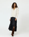 A woman with curly hair is wearing a Suzanne Just Add Love Cashmere Cardigan by Kerri Rosenthal, featuring yellow buttons, over a long, tiered black skirt. She has brown boots on and stands against a plain white background.