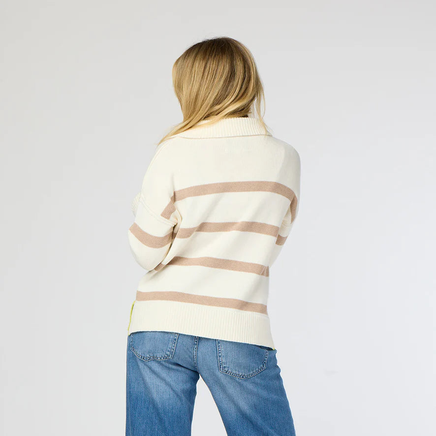 A person with long blonde hair is seen from behind wearing a Kerri Rosenthal Syd Sweater, featuring a cream cotton knit design with tan horizontal stripes, paired with blue jeans against a plain light gray background.