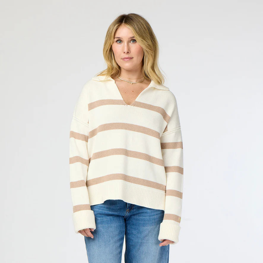 A person with shoulder-length blonde hair poses against a plain background, wearing the Syd Sweater from Kerri Rosenthal, featuring cozy cotton knit fabric with beige horizontal stripes, paired with blue jeans.