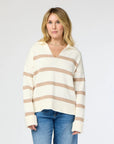 A person with shoulder-length blonde hair poses against a plain background, wearing the Syd Sweater from Kerri Rosenthal, featuring cozy cotton knit fabric with beige horizontal stripes, paired with blue jeans.