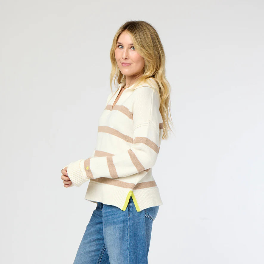 A person with long blonde hair is wearing the Syd Sweater by Kerri Rosenthal, featuring cream and beige stripes with yellow trim at the hem, paired with blue jeans. They are standing sideways and looking at the camera against a plain background.