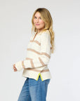 A person with long blonde hair is wearing the Syd Sweater by Kerri Rosenthal, featuring cream and beige stripes with yellow trim at the hem, paired with blue jeans. They are standing sideways and looking at the camera against a plain background.