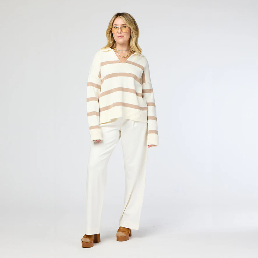 A person wearing the "Syd Sweater," a white and beige striped cashmere piece by Kerri Rosenthal, along with white pants and brown platform shoes, stands against a plain background. They have shoulder-length hair and glasses.