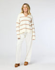 A person wearing the "Syd Sweater," a white and beige striped cashmere piece by Kerri Rosenthal, along with white pants and brown platform shoes, stands against a plain background. They have shoulder-length hair and glasses.