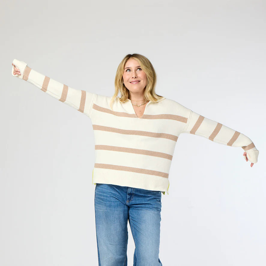 A person wearing the cozy Syd Sweater by Kerri Rosenthal and blue jeans stands with arms outstretched, looking up and smiling against a plain light-colored background.