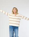 A person wearing the cozy Syd Sweater by Kerri Rosenthal and blue jeans stands with arms outstretched, looking up and smiling against a plain light-colored background.