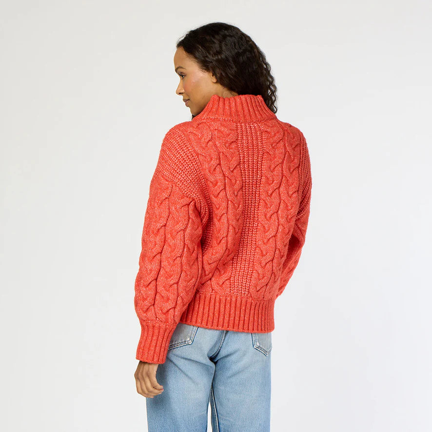 A person with long, wavy hair is wearing a red Troy Cable Sweater from Kerri Rosenthal, crafted from recycled yarn, along with light blue jeans. They are turning slightly to the side with their hand in their pocket against a plain white background.