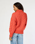 A person with long, wavy hair is wearing a red Troy Cable Sweater from Kerri Rosenthal, crafted from recycled yarn, along with light blue jeans. They are turning slightly to the side with their hand in their pocket against a plain white background.