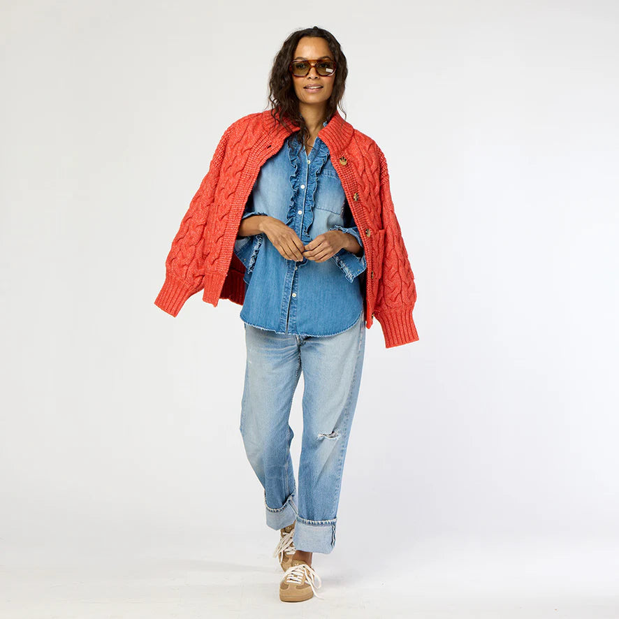 A person wearing sunglasses and a Kerri Rosenthal Troy Cable Sweater crafted from recycled yarn over a blue denim shirt walks confidently. Sporting light blue jeans and beige sneakers, they look stylish and relaxed against a plain white background.