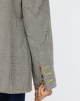 A close-up shot features an individual sporting the gray Workday Plaid Blazer by Kerri Rosenthal, highlighting the sleeve detail. Heart-shaped elbow patches seamlessly integrate into the plaid design, while three brass buttons adorn the cuff. The person's arm rests at their side over dark denim pants against a simple backdrop.