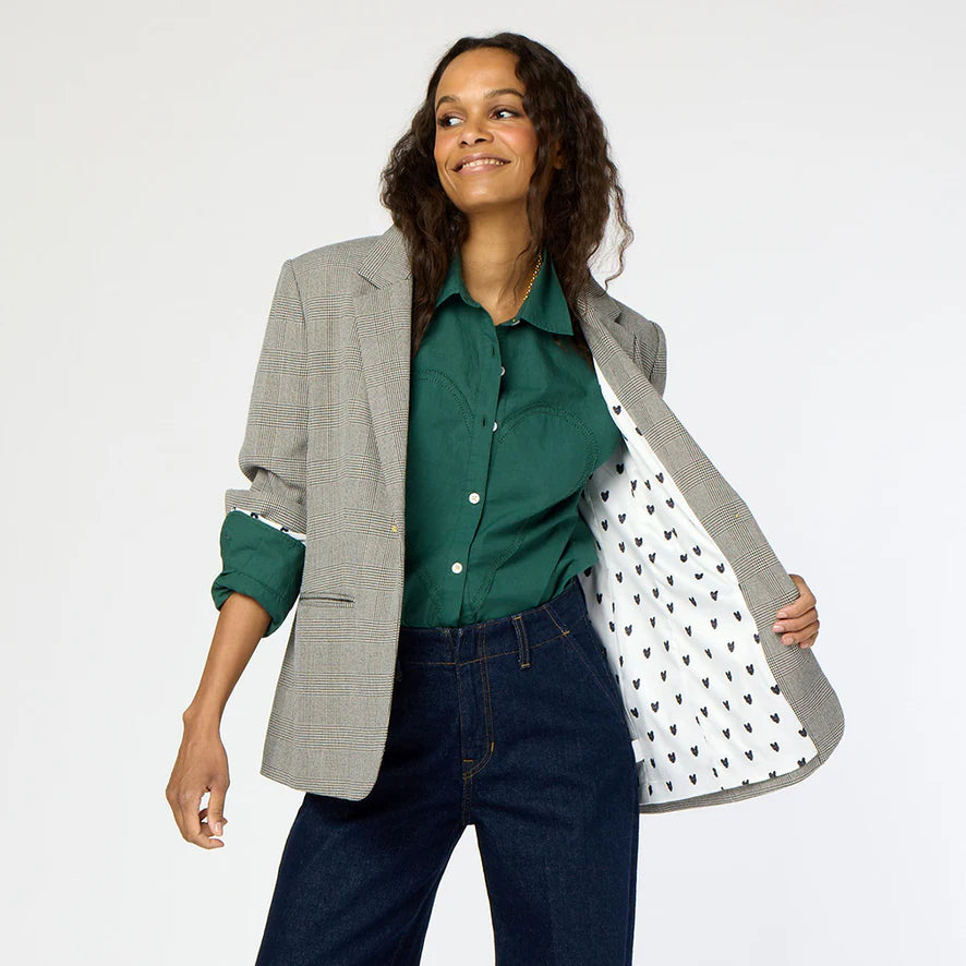 A person wearing a green shirt, a Kerri Rosenthal Workday Plaid Blazer with heart elbow patches, and dark blue jeans smiles while holding one side of the blazer open to reveal the patterned lining. Their long, wavy hair flows with confidence against a plain background.