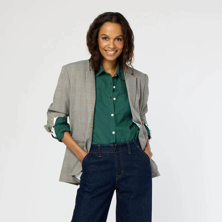 Wearing her long hair down, a person beams while dressed in Kerri Rosenthal's Workday Plaid Blazer featuring heart elbow patches over a green shirt and blue jeans. With hands in their pockets, they embrace the day against a plain background.