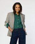 Wearing her long hair down, a person beams while dressed in Kerri Rosenthal's Workday Plaid Blazer featuring heart elbow patches over a green shirt and blue jeans. With hands in their pockets, they embrace the day against a plain background.