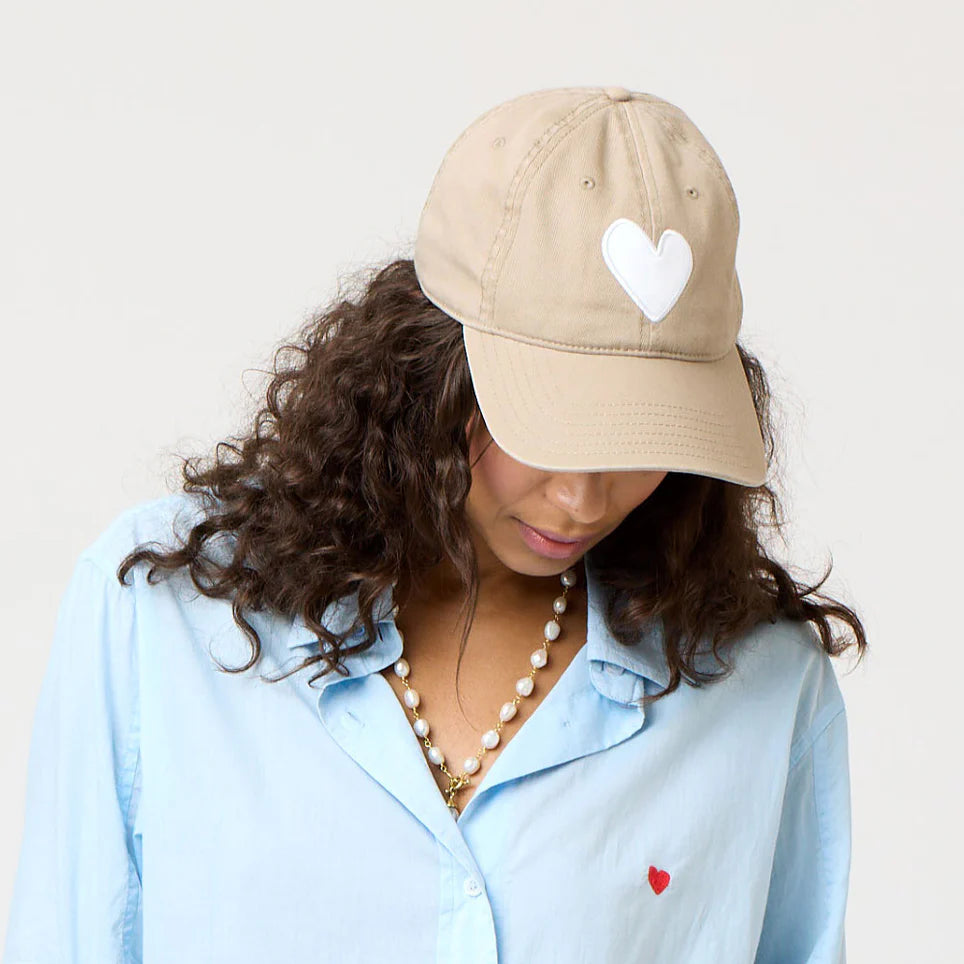 Wearing a Kerri Rosenthal KR Imperfect Heart Hat with a white heart emblem, a person looks down, revealing curly hair under the beige cap. They sport a light blue shirt adorned with a red heart and wear a pearl necklace against a plain backdrop—an exclusive collector&#39;s style.