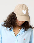 Wearing a Kerri Rosenthal KR Imperfect Heart Hat with a white heart emblem, a person looks down, revealing curly hair under the beige cap. They sport a light blue shirt adorned with a red heart and wear a pearl necklace against a plain backdrop—an exclusive collector's style.