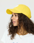 A woman with long curly hair wears a yellow KR Imperfect Heart Hat by Kerri Rosenthal, featuring an embroidered tonal heart patch, with a white t-shirt and layered gold necklaces, standing against a plain light background.