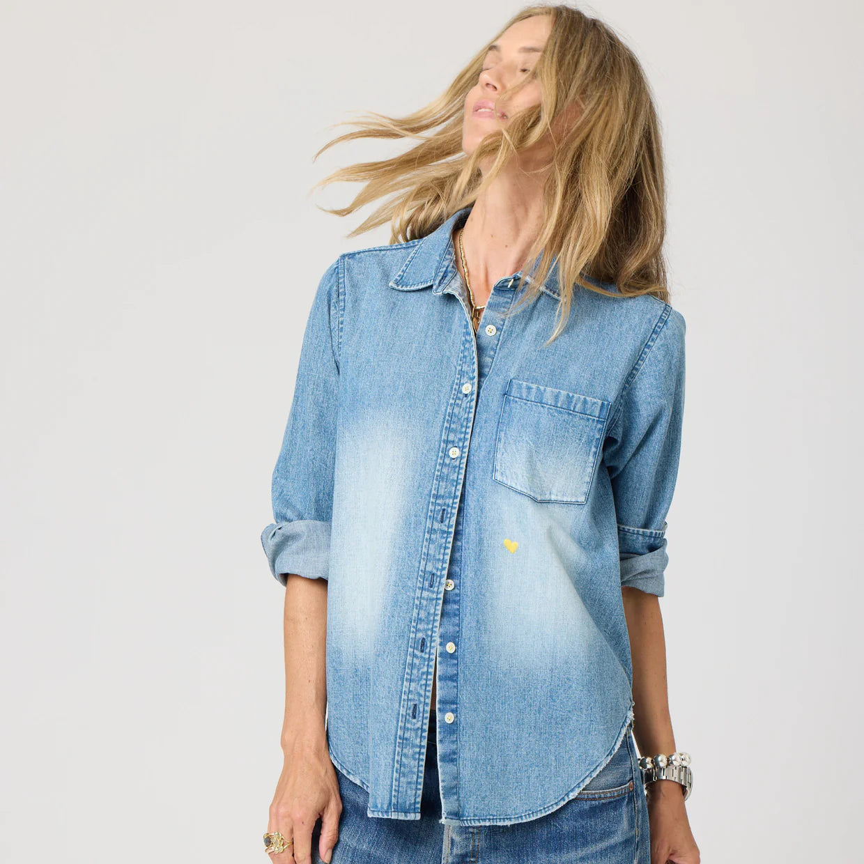 A person with long blonde hair wears the Kerri Rosenthal Denim Shirt Lightweight, featuring rolled-up sleeves and faded front patches, turned slightly to the side against a plain light background.