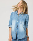 A person with long blonde hair wears the Kerri Rosenthal Denim Shirt Lightweight, featuring rolled-up sleeves and faded front patches, turned slightly to the side against a plain light background.