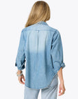 A woman with long blonde hair wears the Kerri Rosenthal Denim Shirt Lightweight, featuring rolled-up sleeves and embroidered heart elbow details, standing with her back to the camera against a plain background.