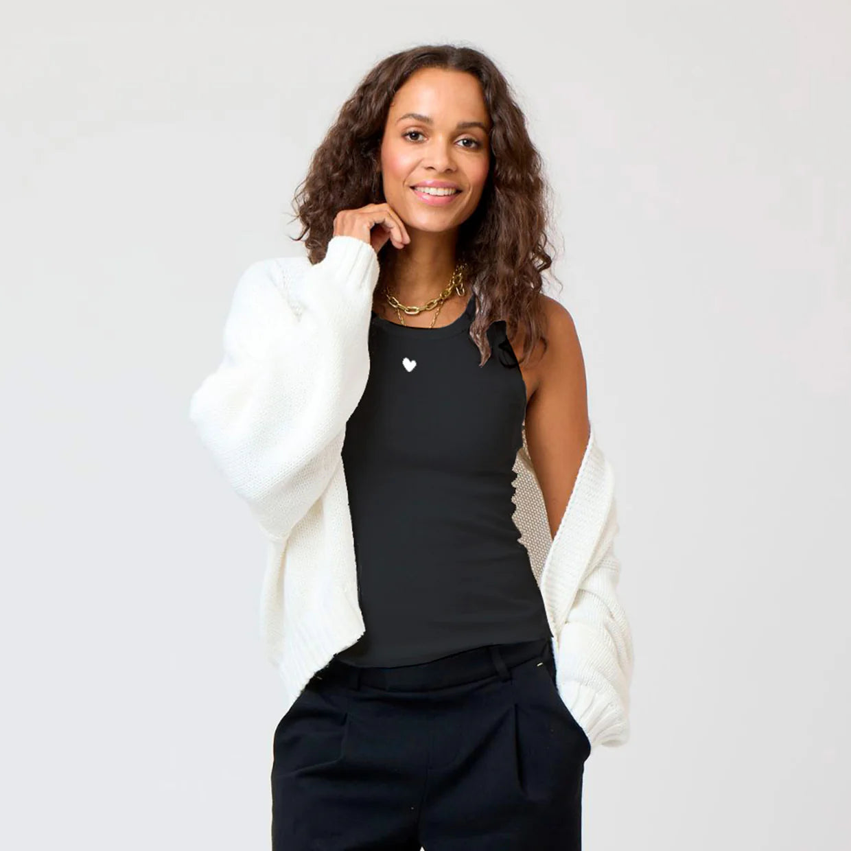 A woman with curly hair smiles while wearing the KR Tank by Kerri Rosenthal, a black cotton tank with an embroidered heart, paired with black pants and a white cardigan draped over her shoulders against a plain background.