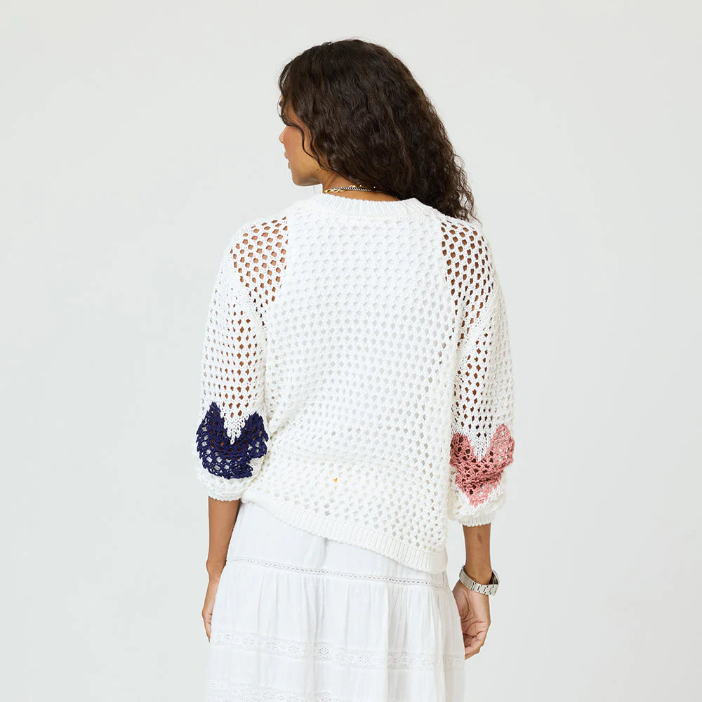 A woman with long curly hair, seen from behind, wears the Kerri Rosenthal Catch Open Stitch Sweater with navy and pink sleeve accents and a white skirt against a plain, light background.