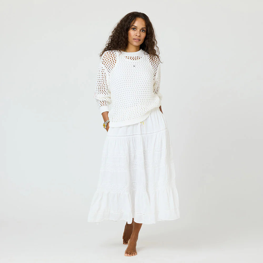 A woman with curly hair stands barefoot against a plain background, wearing the Kerri Rosenthal Catch Open Stitch Sweater—a white, 100% cotton crew neck—paired with a flowing white skirt.