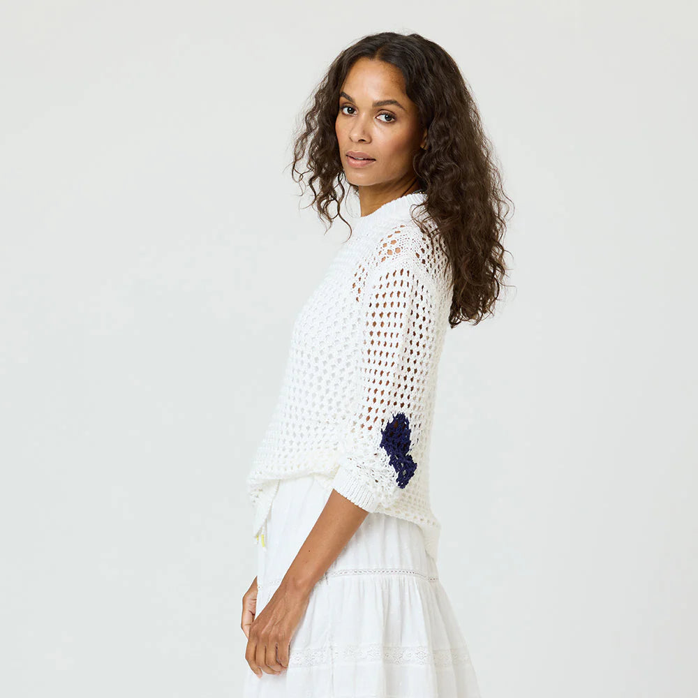 A woman with long curly hair models the Kerri Rosenthal Catch Open Stitch Sweater, featuring blue sleeve detailing, paired with a white skirt. She stands sideways against a plain light background, looking at the camera.