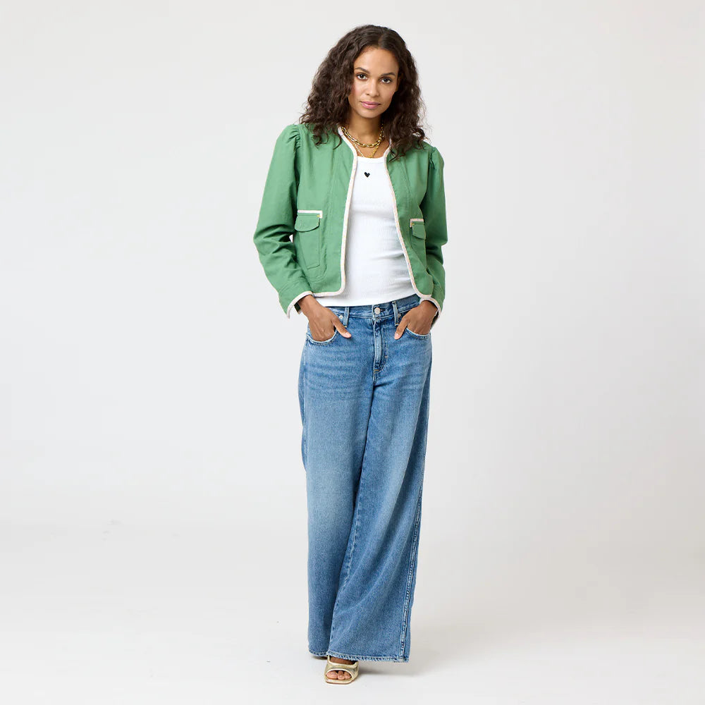 A person with long curly hair stands in a green Morgan Beau-Lero jacket by Kerri Rosenthal, a white shirt, and wide-leg jeans. They have their hands in their pockets on a light gray background and are wearing sandals.