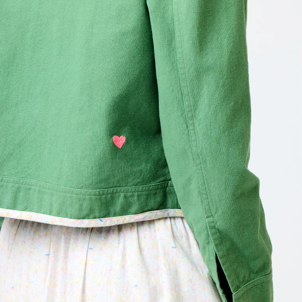 A person wears a Kerri Rosenthal Morgan Beau-Lero green jacket with a small pink heart embroidery, paired with a white skirt featuring Sweetie Pie print&#39;s light, multicolored pattern against a plain white backdrop.