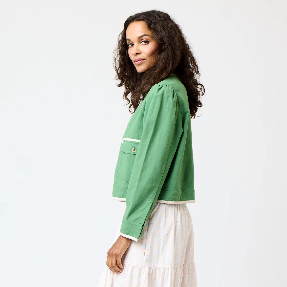 A woman with curly hair poses sideways, wearing the Morgan Beau-Lero Jacket by Kerri Rosenthal over a white dress against a plain light background.