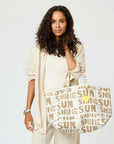 A woman with curly hair, dressed in cream, carries the KR Sunshine Tote by Kerri Rosenthal—featuring "SUNSHINE" in bold letters. She stands against a plain white background, looking into the camera.