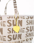 The KR Sunshine Tote by Kerri Rosenthal is a cotton canvas bag with "SUN" and "SHINE" in brown block letters, plus a yellow heart tag on the handle. Inspired by Kerri’s Sunshine Artwork.