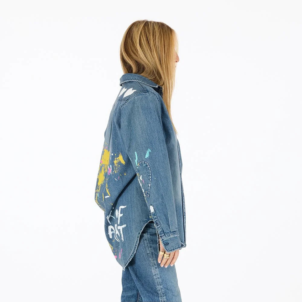 A person with long blonde hair stands sideways in a relaxed fit Kerri Rosenthal Mia Studio Shirt and jeans. The denim shirt features colorful painted designs and text on the back, set against a plain white background.