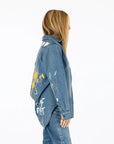 A person with long blonde hair stands sideways in a relaxed fit Kerri Rosenthal Mia Studio Shirt and jeans. The denim shirt features colorful painted designs and text on the back, set against a plain white background.