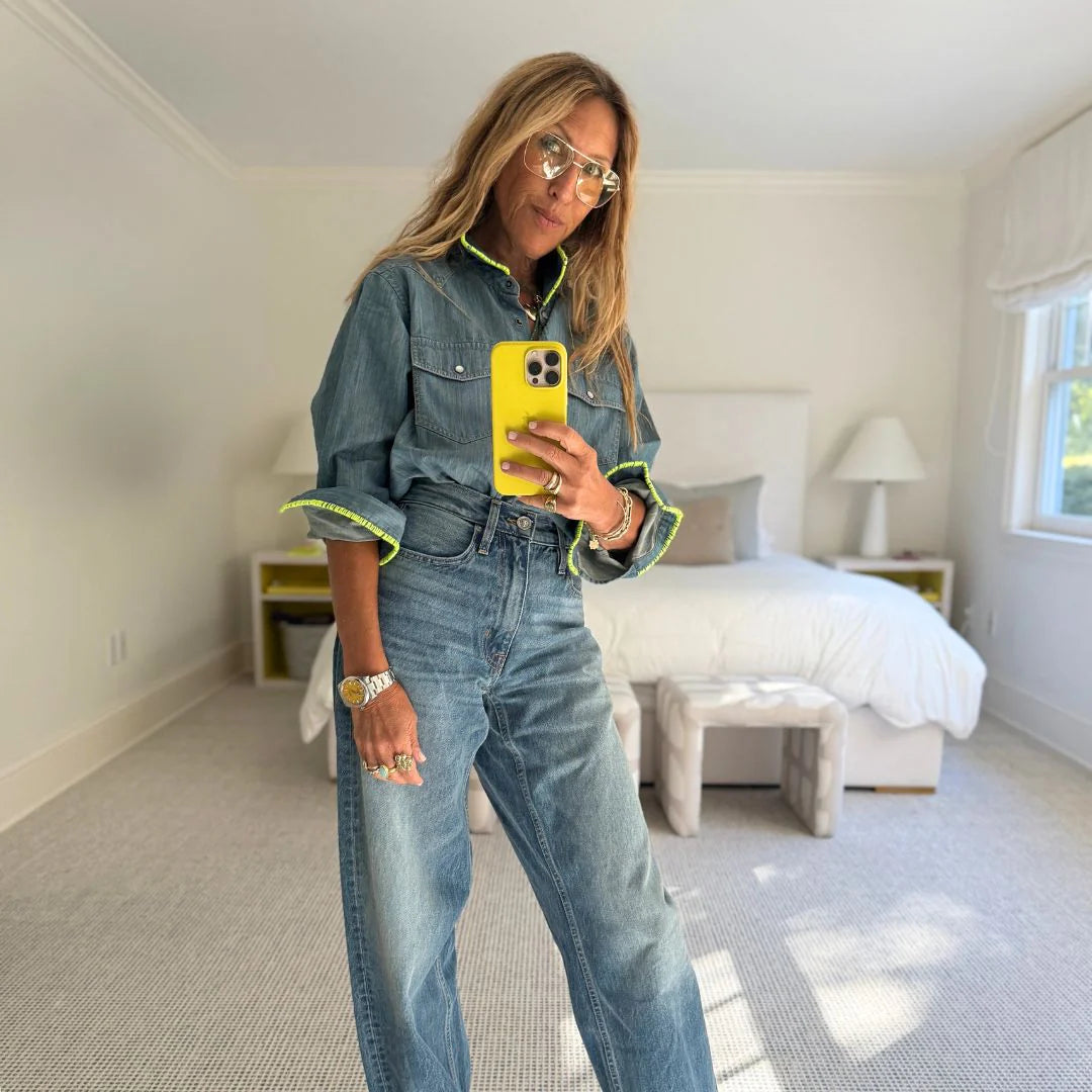 A woman with long hair and glasses takes a mirror selfie in a bright, modern bedroom, wearing the Kerri Rosenthal Pia Denim Western Shirt and high-waisted jeans.