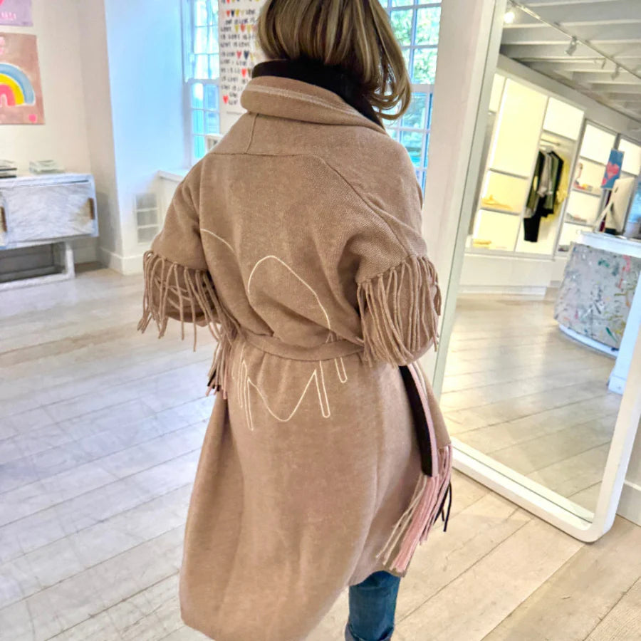 A person models the Kerri Rosenthal Cali Fringe Cardigan Coat indoors, facing away in a bright room with large windows, white walls, wooden floors, and vibrant displays of artwork and clothing.